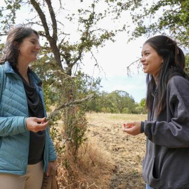 A student and professor, who is holding a tree branch, are smiling and looking at each other while outside